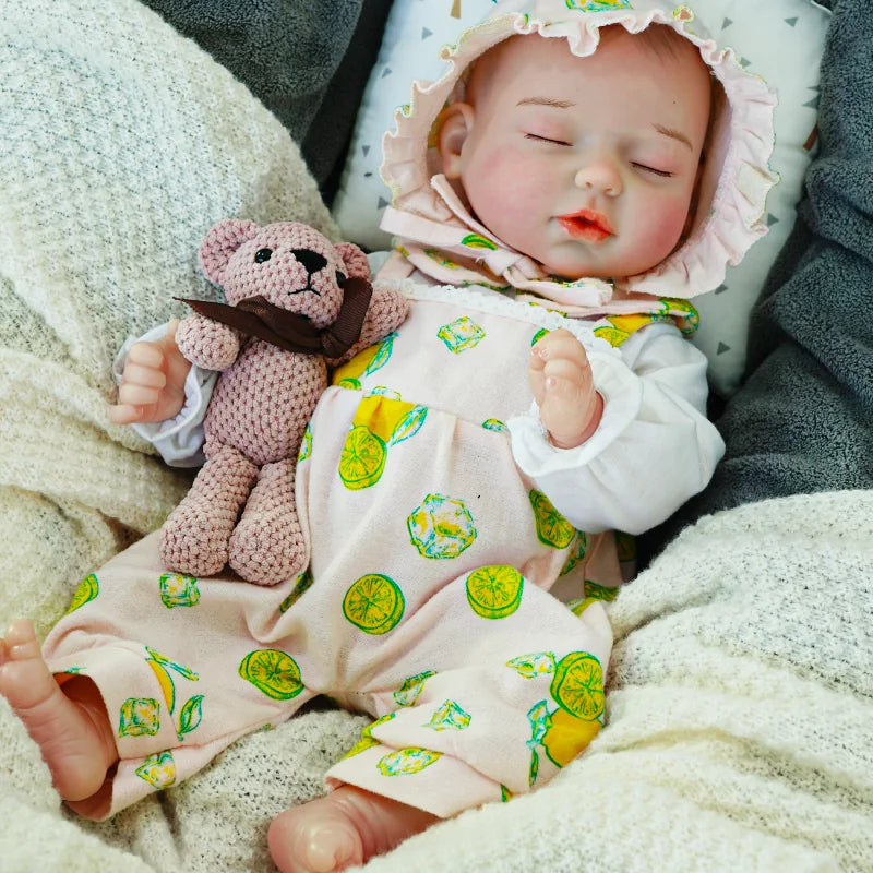 A peaceful reborn baby doll with a bonnet, eyes closed, wearing a white and green patterned outfit, and holding a small knitted bear.