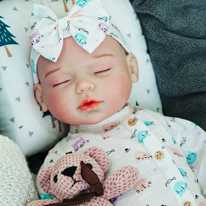 The reborn baby doll resting with a serene expression, dressed in a blue-patterned onesie and headband, holding a knitted bear.