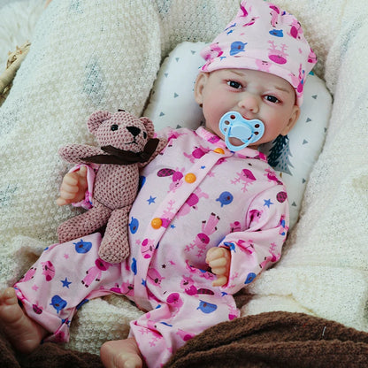 The reborn baby doll with a pacifier, wearing a pink outfit with animal prints, holding a small teddy bear, and resting on a cushion.