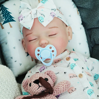 The reborn baby doll with a pacifier, peacefully sleeping, wearing a light onesie with a bow headband and holding a small teddy bear.