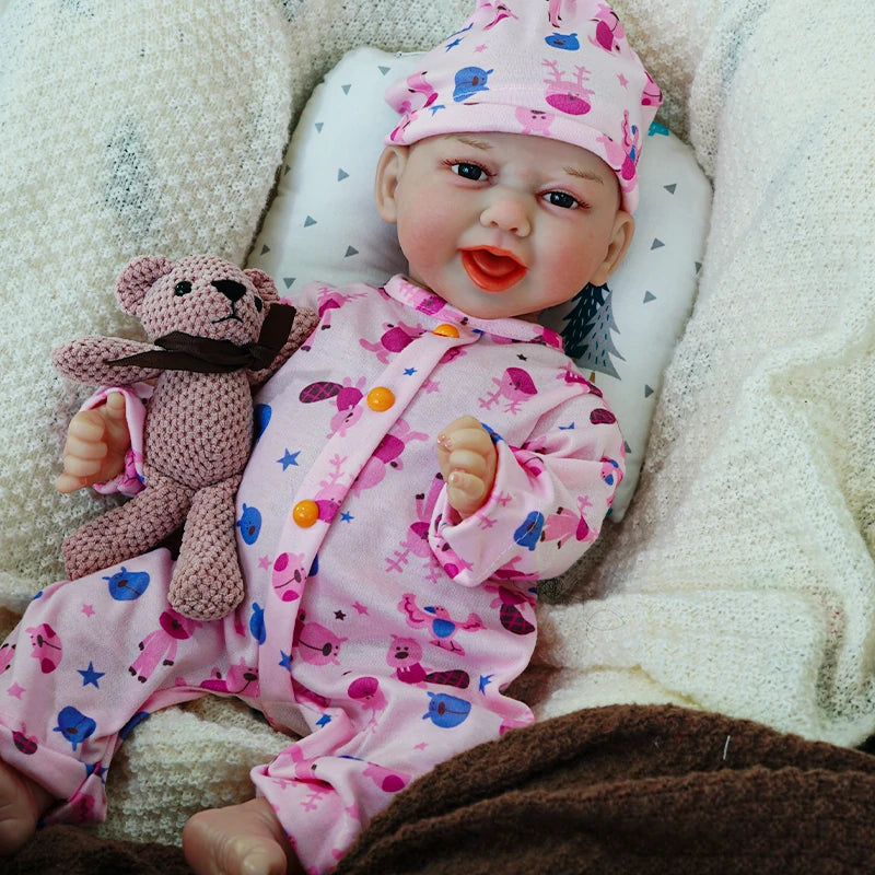 The reborn baby doll sitting up, smiling, dressed in a pink animal print onesie, with a knitted bear.