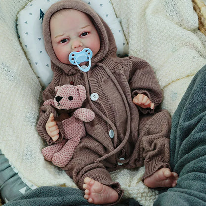 The reborn baby doll lying down with a pacifier, dressed in a brown hooded onesie, cuddling with a knitted teddy bear.