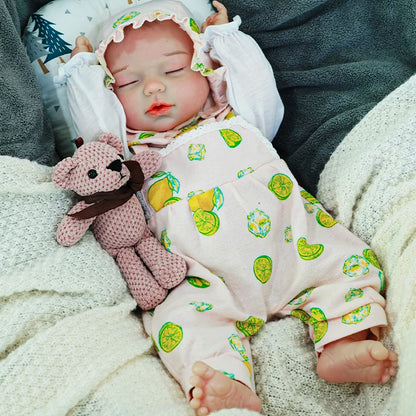 A serene reborn baby doll wearing a bonnet and a green onesie, holding a knitted bear, and resting on a blanket.