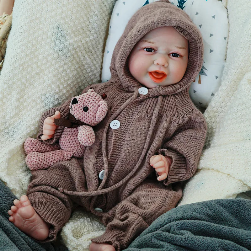 The reborn baby doll smiling brightly, dressed in a brown knit outfit with a hood, holding a soft teddy bear.