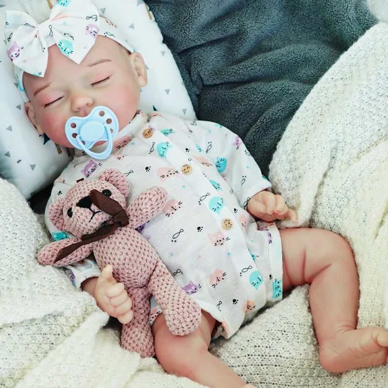 A peaceful reborn baby doll with a pacifier, dressed in a blue-patterned onesie with a bow headband, lying down with its teddy bear.