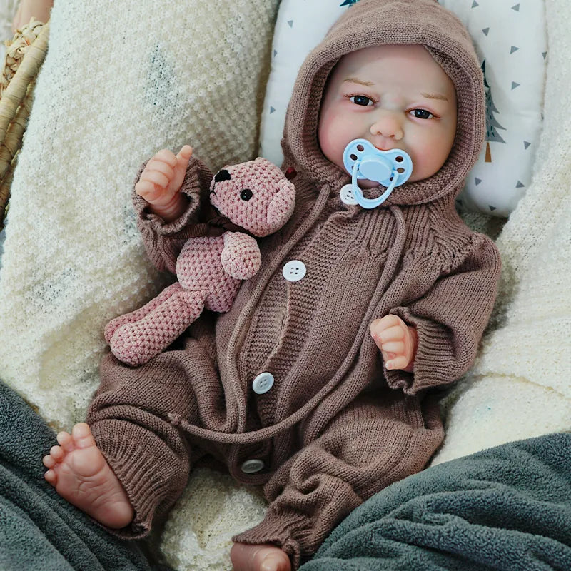 The reborn baby doll sitting up with a pacifier, dressed in a cozy brown knit onesie, holding a knitted teddy bear.