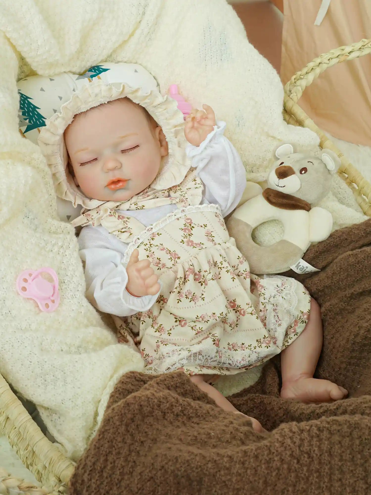 Reborn baby doll in a floral bonnet and dress, sleeping peacefully in a basket, cuddled by plush blankets and a teddy bear, exuding a serene nursery atmosphere.