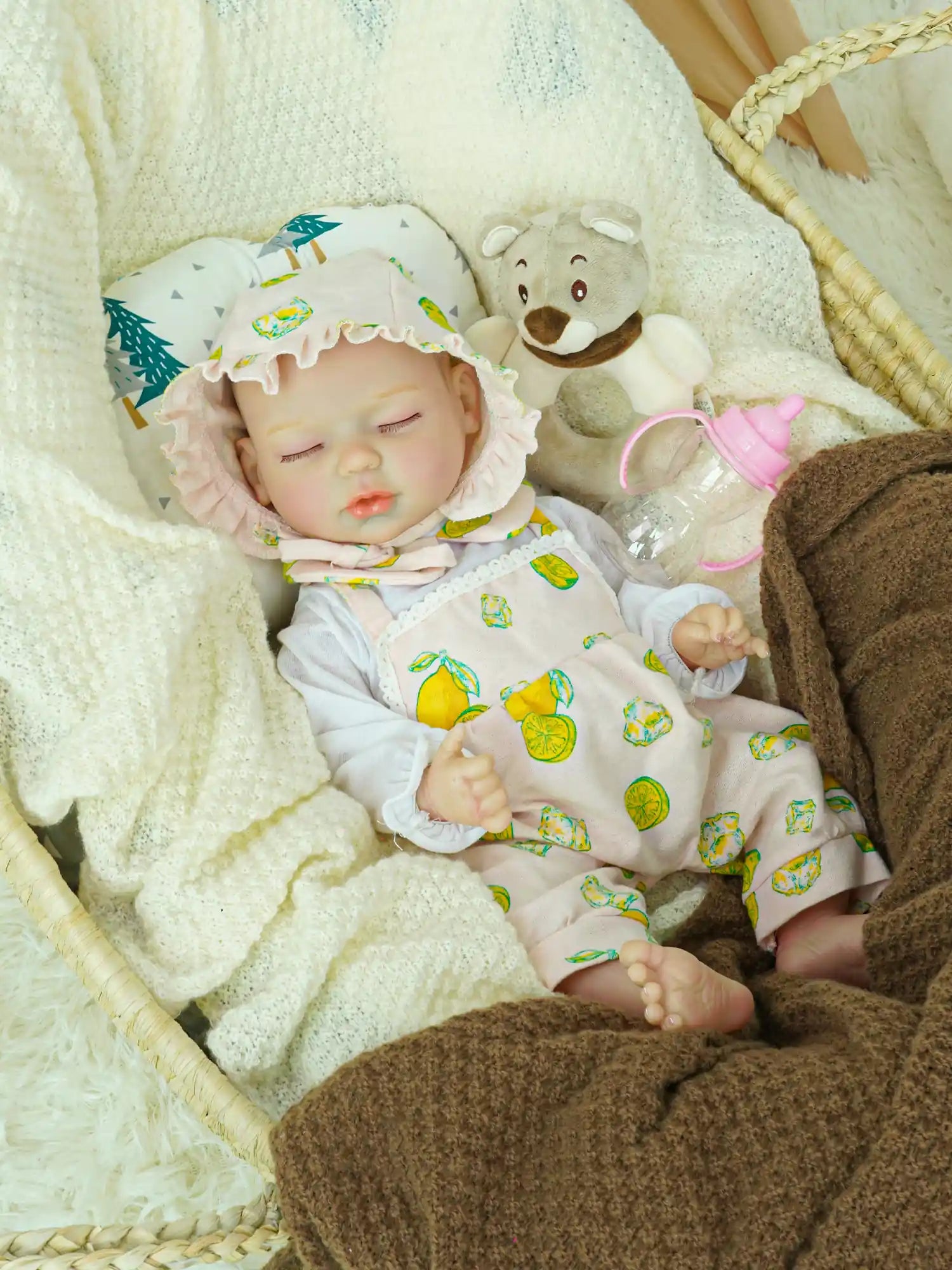 A serene reborn baby doll adorned in a pink outfit with lemon patterns and a matching bonnet, lying in a basket filled with soft blankets. The scene is completed with a plush teddy bear and a baby bottle, evoking a nurturing environment.
