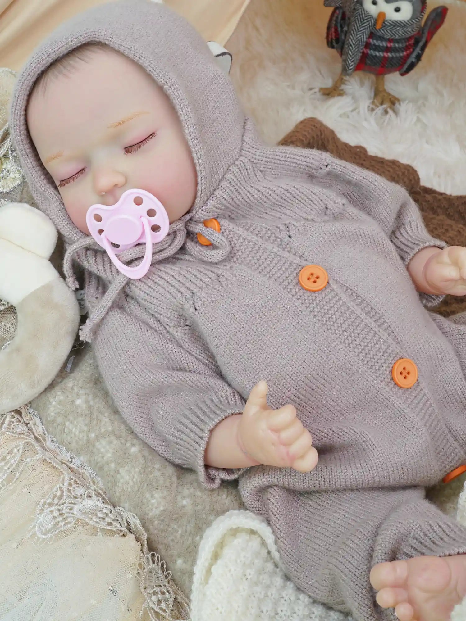 A close-up of a reborn doll toy in a cozy lilac knitted outfit with a hood, pacifier in mouth, lying on a plush white surface surrounded by soft toys