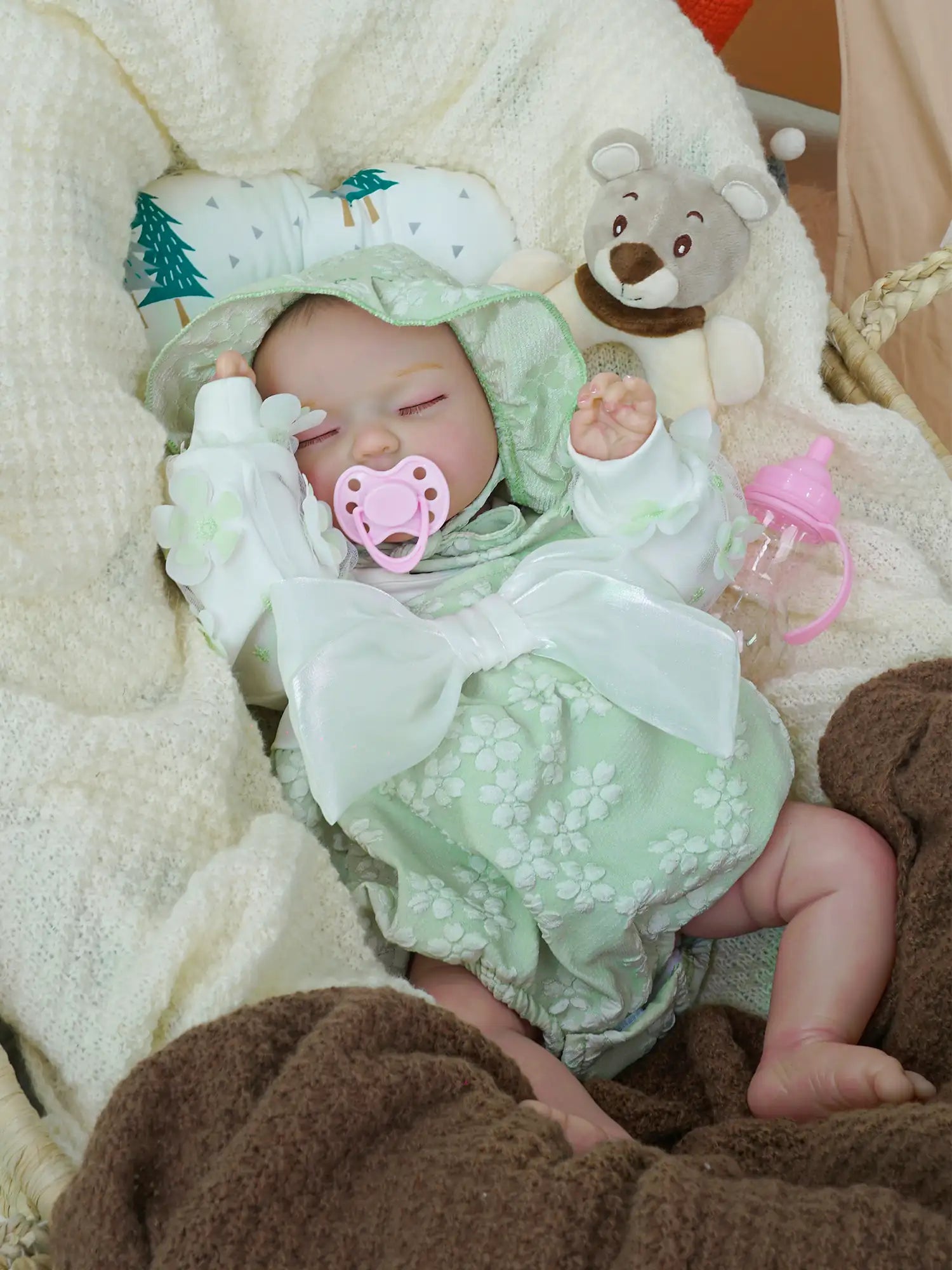 Reborn baby doll in a pale green floral outfit with a matching hood, sleeping comfortably in a wicker basket. The doll is surrounded by soft white and brown blankets, with a teddy bear and a pink bottle positioned nearby.