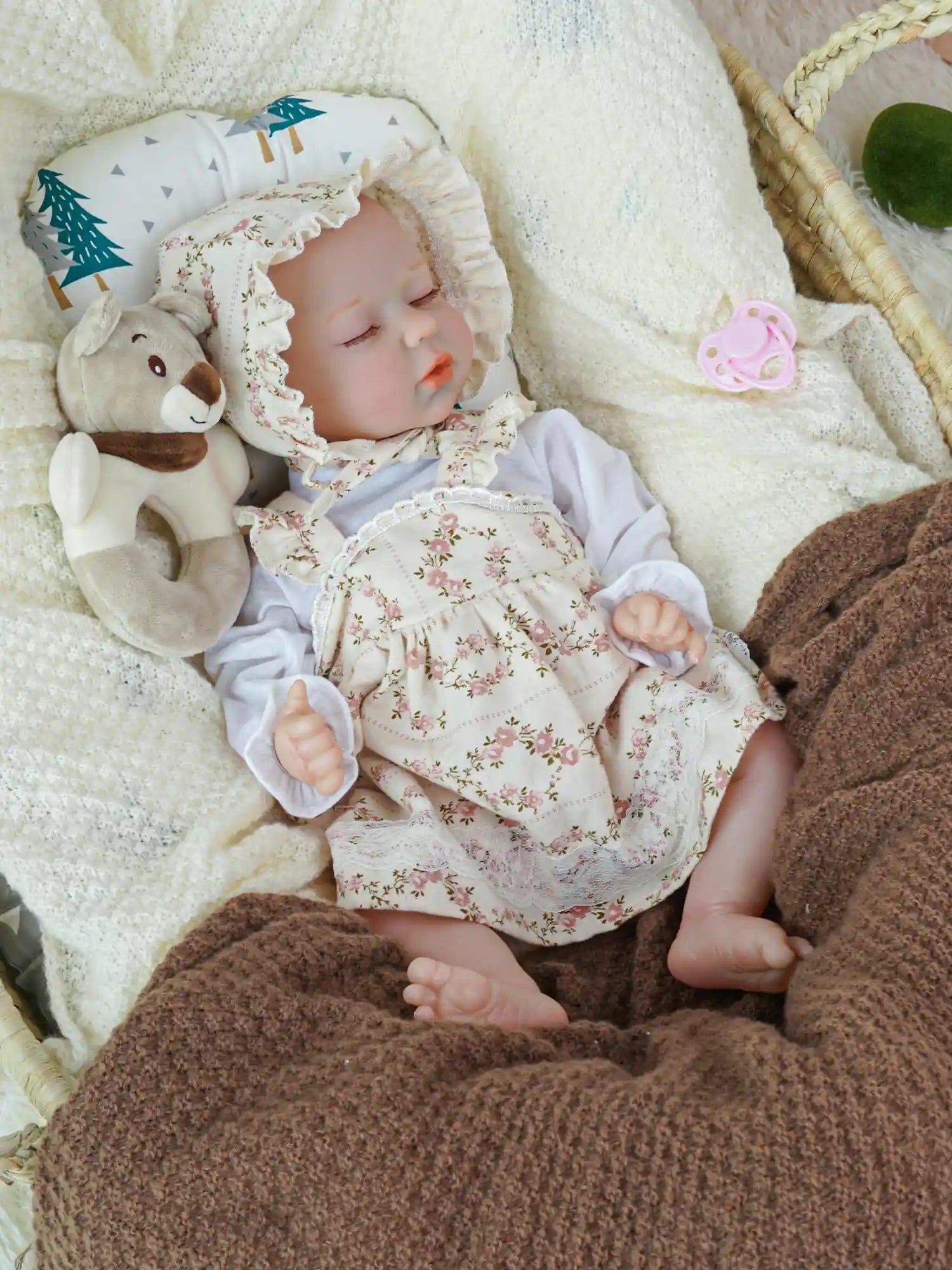 A reborn baby doll with a floral bonnet and matching dress, peacefully sleeping in a woven basket, nestled in a cream-colored blanket. A pink pacifier is in its mouth, and a plush teddy bear lies beside it.