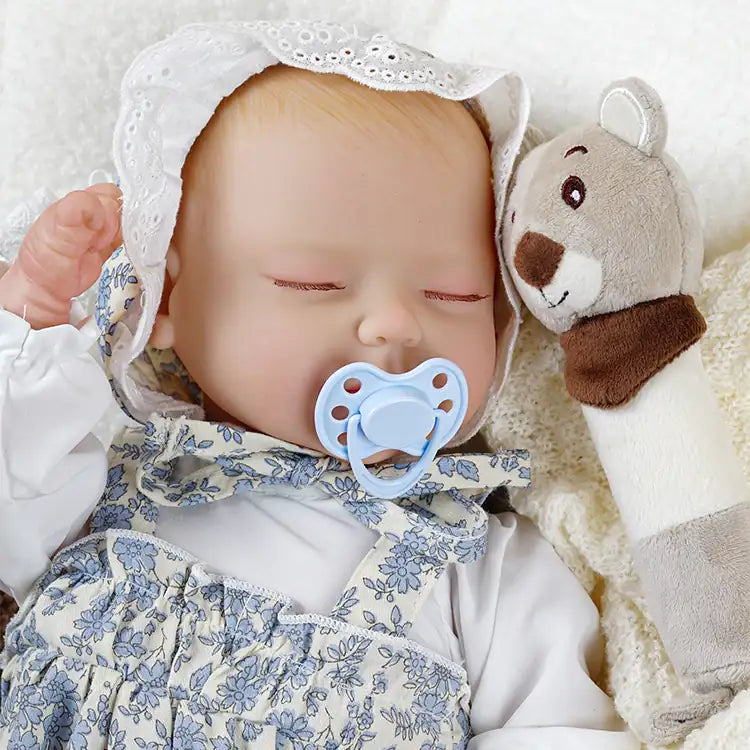 A lifelike reborn doll sleeping peacefully with a blue pacifier in its mouth. The doll is wearing a white and blue floral outfit with a lace bonnet and is holding a plush toy