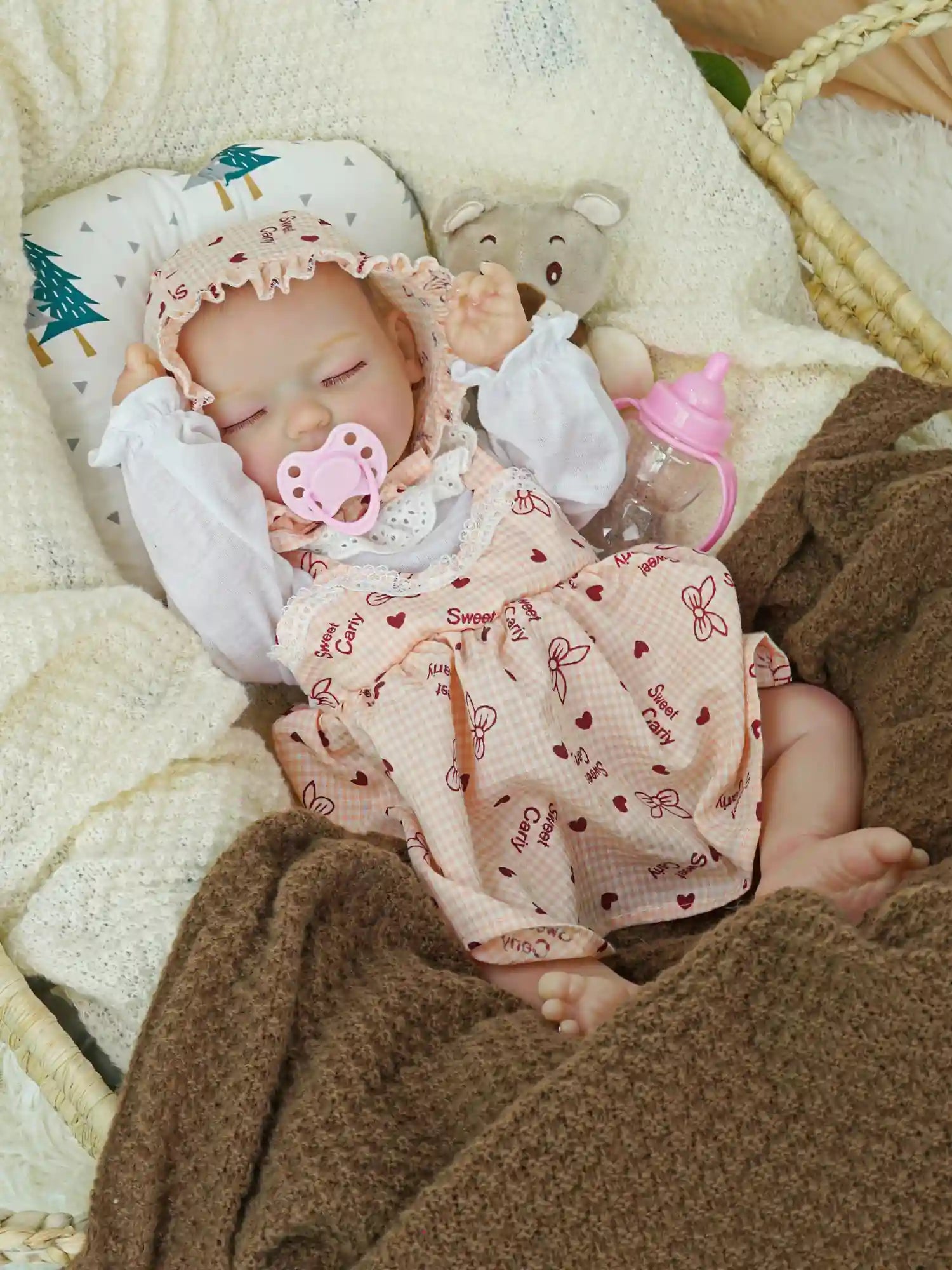 This image shows a reborn doll resting on a layered blanket, wearing a pink bonnet and a dress with red hearts, with a pink baby bottle and a gray teddy bear nearby.
