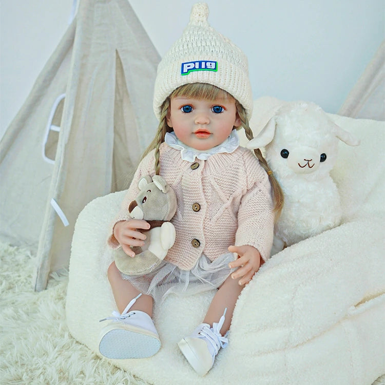 A reborn toddler doll sitting next to a teddy bear, wearing a pink coat and white shoes.