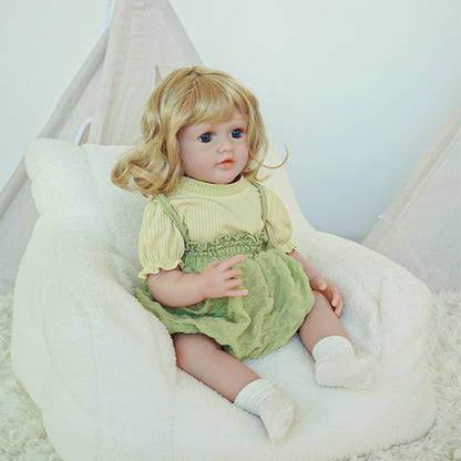 Reborn toddler doll sitting in a plush chair, dressed in a green dress with white socks, looking slightly to the side.