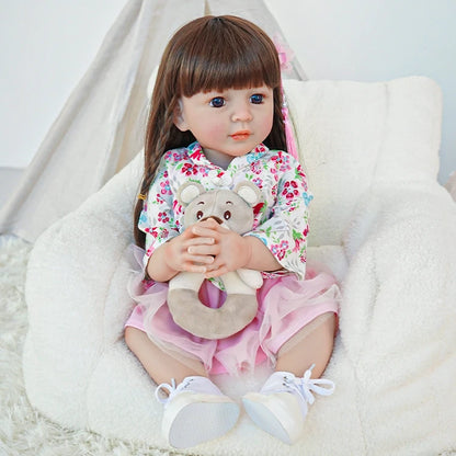 Reborn toddler doll sitting on a plush surface, dressed in a floral top and pink skirt, holding a teddy bear.