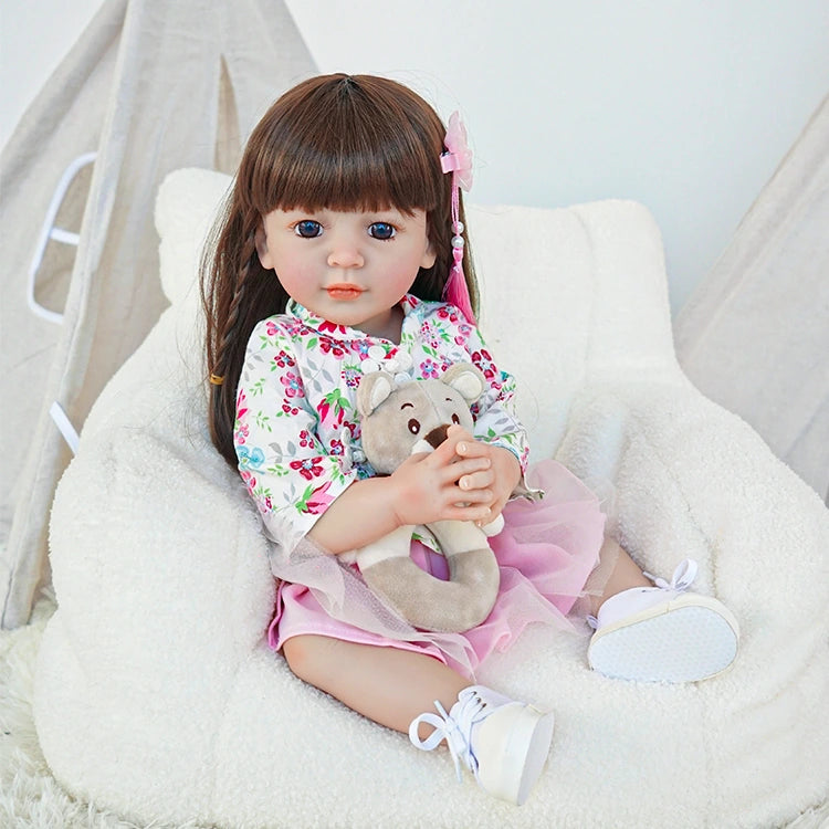 Reborn toddler doll sitting in a soft chair, dressed in a floral top and pink skirt, holding a small teddy bear.