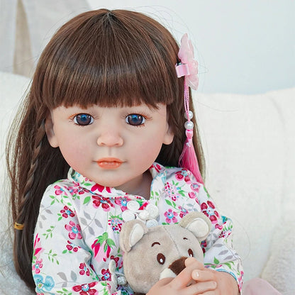 Close-up of reborn toddler doll with pink hair ribbons, wearing a floral pink outfit, holding a teddy bear.