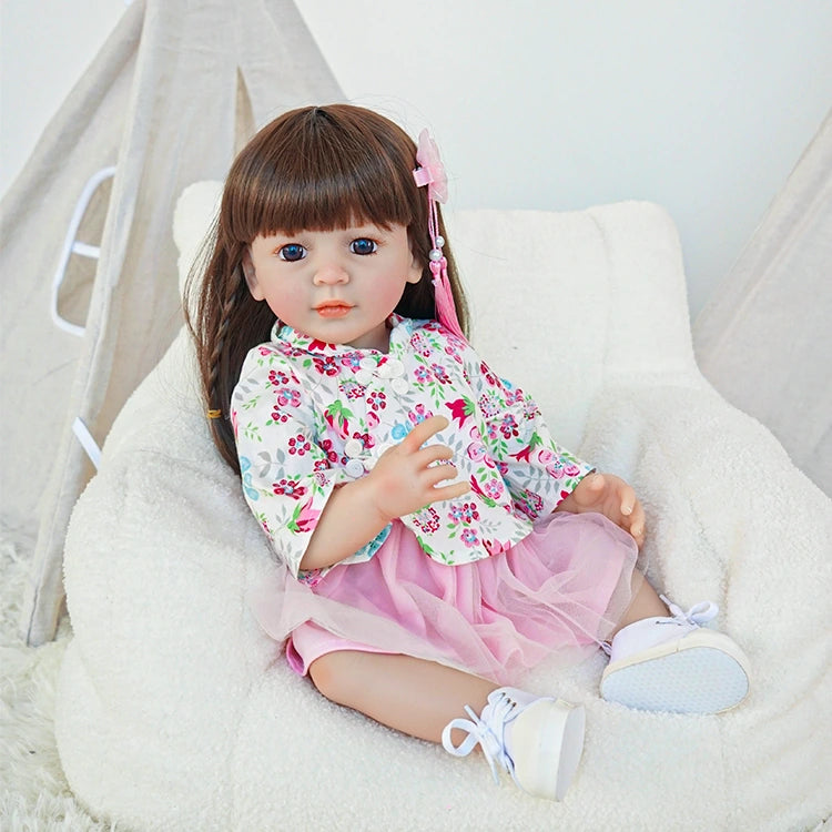 Reborn toddler doll sitting on a plush chair, dressed in a floral pink top and skirt, holding a small teddy bear.