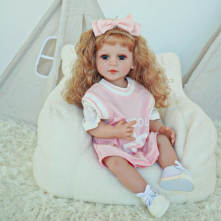 Reborn toddler doll sitting on a plush chair, wearing a pink dress with a large bow in her curly hair.