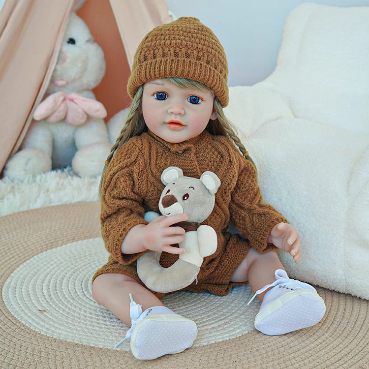 Reborn toddler doll wearing a brown knit outfit and matching hat, holding a small teddy bear while sitting on a rug.