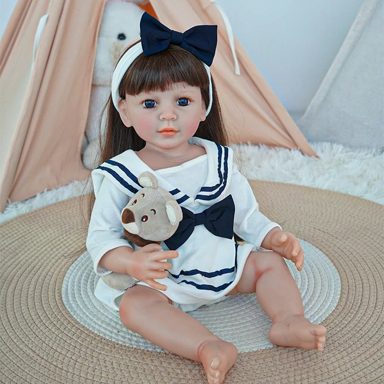 
Reborn toddler doll sitting on a rug, wearing a sailor-style outfit with a large navy bow, holding a plush teddy bear.