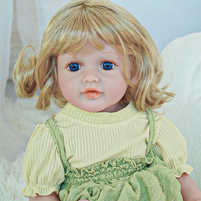 Reborn toddler doll sitting on a soft surface, dressed in a light green dress with a striped top, looking forward with a gentle expression.