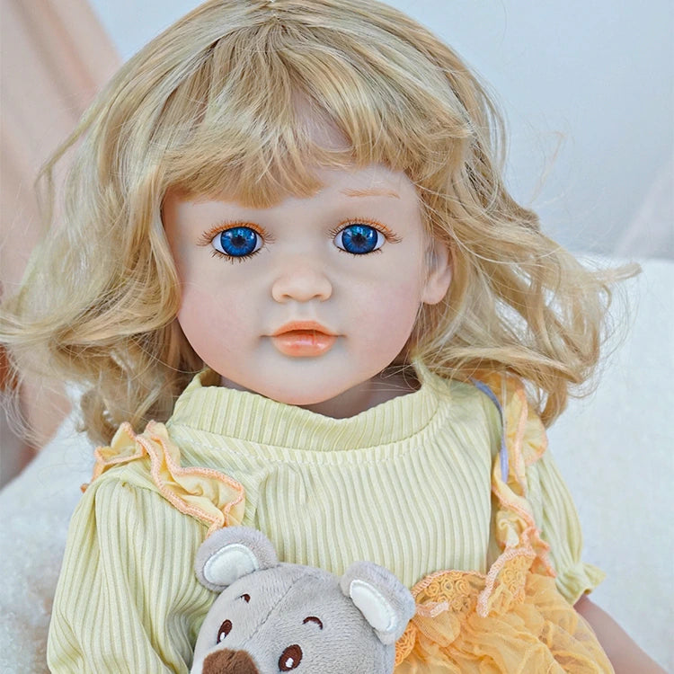  Reborn toddler doll sitting on a soft surface, dressed in a yellow dress with floral patterns, holding a small teddy bear, looking forward.