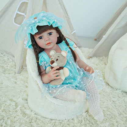 Reborn toddler doll sitting on a soft surface, dressed in a light blue dress with a matching bonnet, holding a teddy bear.