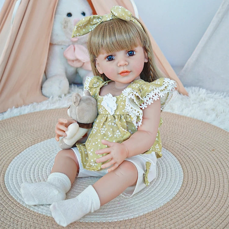 Reborn toddler doll sitting on a soft rug, dressed in a green floral dress with lace details, holding a small teddy bear.