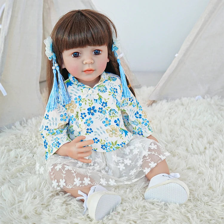 Reborn toddler doll sitting on a soft rug, dressed in a floral blue and white outfit with white shoes, looking forward.