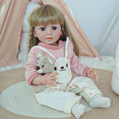 Reborn toddler doll sitting on a soft rug, dressed in a pink sweater and white overalls with a bunny face, holding a small teddy bear.