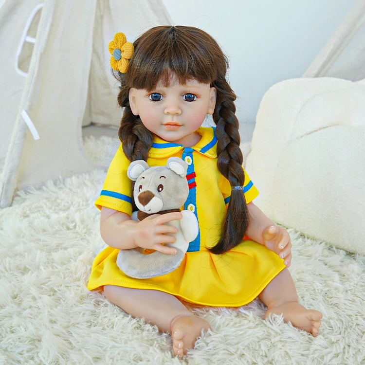 Reborn toddler doll sitting on a soft rug, dressed in a bright yellow dress with blue accents, holding a small teddy bear.