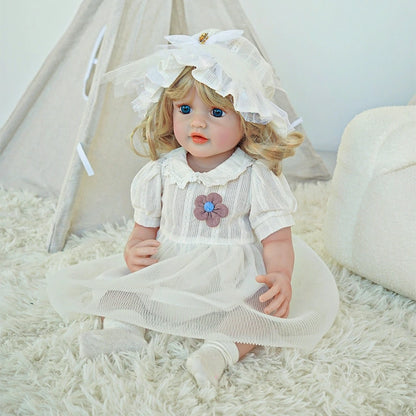 Reborn toddler doll sitting on a soft rug, dressed in a white dress with a floral brooch and matching bonnet.