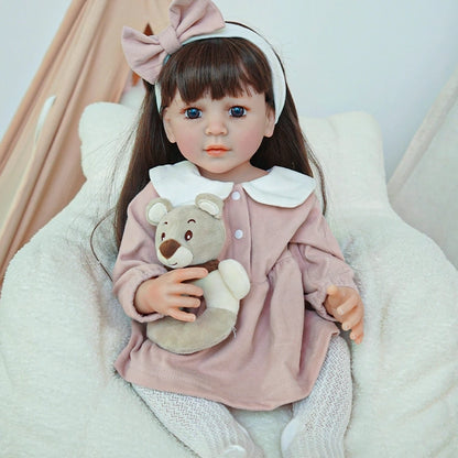 Reborn toddler doll sitting on a soft surface, dressed in a pink outfit with a large bow and holding a small teddy bear.