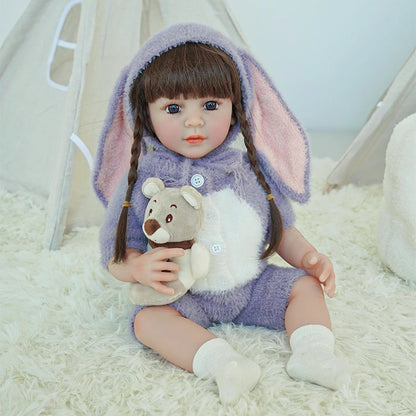 Reborn toddler doll sitting on a soft rug, dressed in a purple bunny-themed outfit with long bunny ears, holding a small teddy bear.