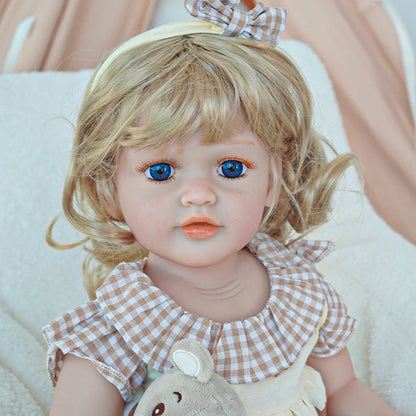 Reborn toddler doll sitting on a soft rug, dressed in a pink sweater and white overalls with a bunny face, holding a small teddy bear.