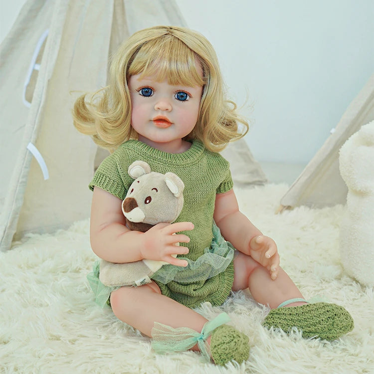 Reborn toddler doll holding a small teddy bear, wearing a green knit outfit, sitting on a soft surface.