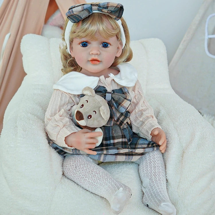 Reborn toddler doll sitting on a soft chair, dressed in a plaid dress with a large white collar, holding a small teddy bear.