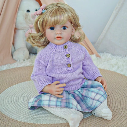 Reborn toddler doll sitting on a soft rug, dressed in a purple sweater and plaid pants, looking forward.