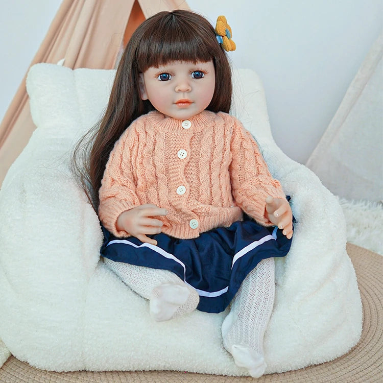 Reborn toddler doll sitting on a soft chair, dressed in a peach knit sweater and navy blue skirt, with white socks.