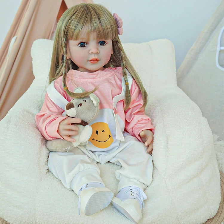 Reborn toddler doll sitting in a soft chair, dressed in a pink top and white overalls, holding a small teddy bear.