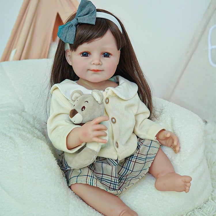 Reborn toddler doll sitting on a soft chair, dressed in a cream cardigan and plaid skirt, holding a small teddy bear.