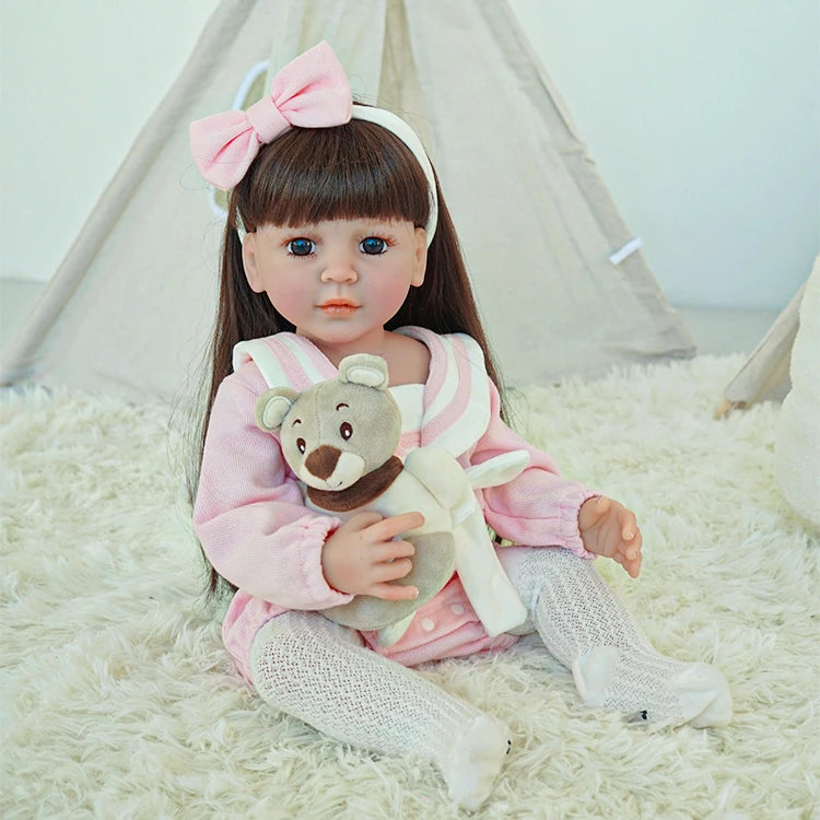 Reborn toddler doll sitting on a soft surface, dressed in a pink outfit with a white bow, holding a small teddy bear.