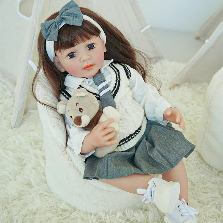 Reborn toddler doll sitting on a soft chair, dressed in a school uniform with a large gray bow, holding a teddy bear.