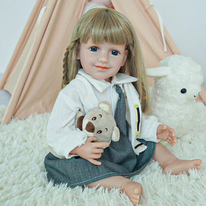 Reborn toddler doll sitting on a soft surface, wearing a white shirt, gray skirt, and holding a small teddy bear.