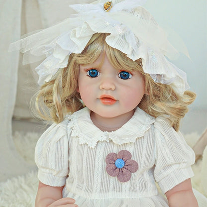 Reborn toddler doll sitting on a rug, wearing a white bonnet and dress with floral details, looking forward.