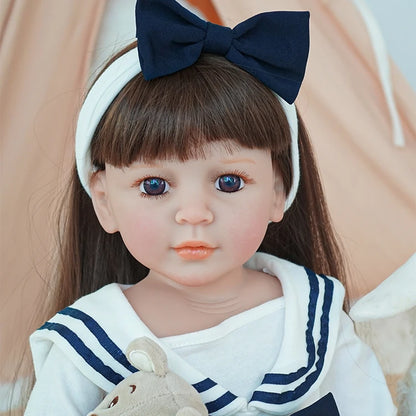 Close-up of reborn toddler doll in a sailor dress with a navy blue bow on her head, holding a small teddy bear, looking directly forward.