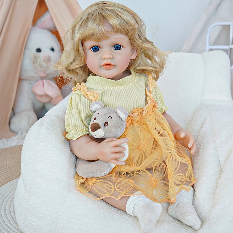 Reborn toddler doll sitting on a plush chair, wearing a yellow dress with floral designs, holding a teddy bear, looking gently to the side.