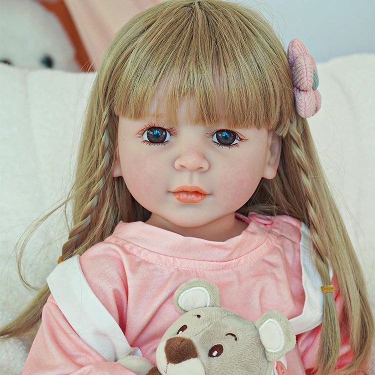 Close-up of reborn toddler doll with long blonde hair and a pink flower clip, wearing a pink top and white overalls.